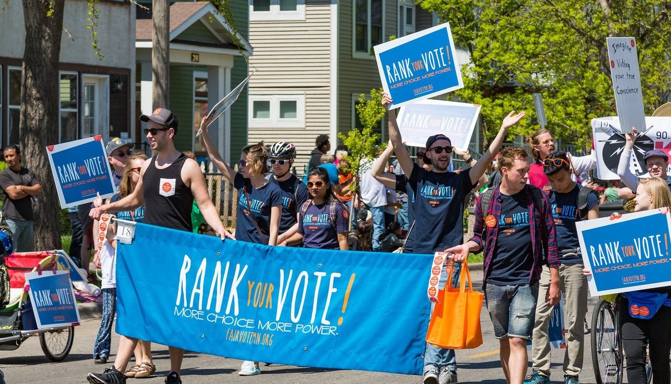 people marching in a parade with various banners and signs that state "Rank Your vote! More Choice. More Power." in support of ranked choice voting